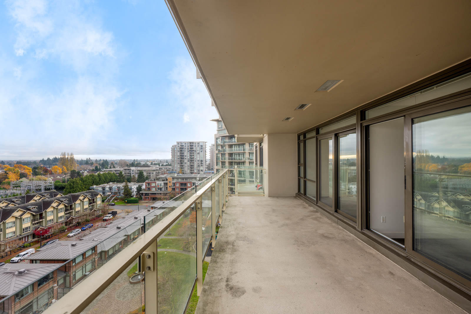 A spacious balcony with glass railing, adjacent to large sliding glass doors, overlooks a cityscape with mid-rise buildings and trees under a partly cloudy sky.