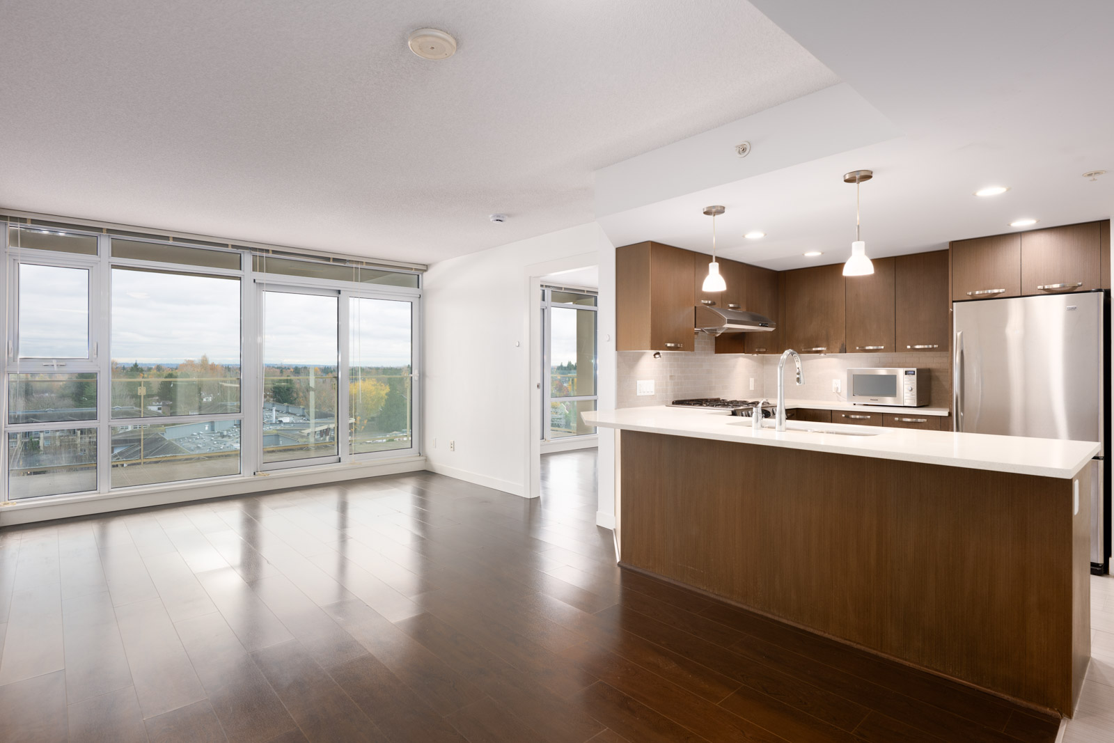 Modern open-concept apartment with large windows, hardwood floors, and a kitchen featuring dark cabinets, pendant lights, and stainless steel appliances.