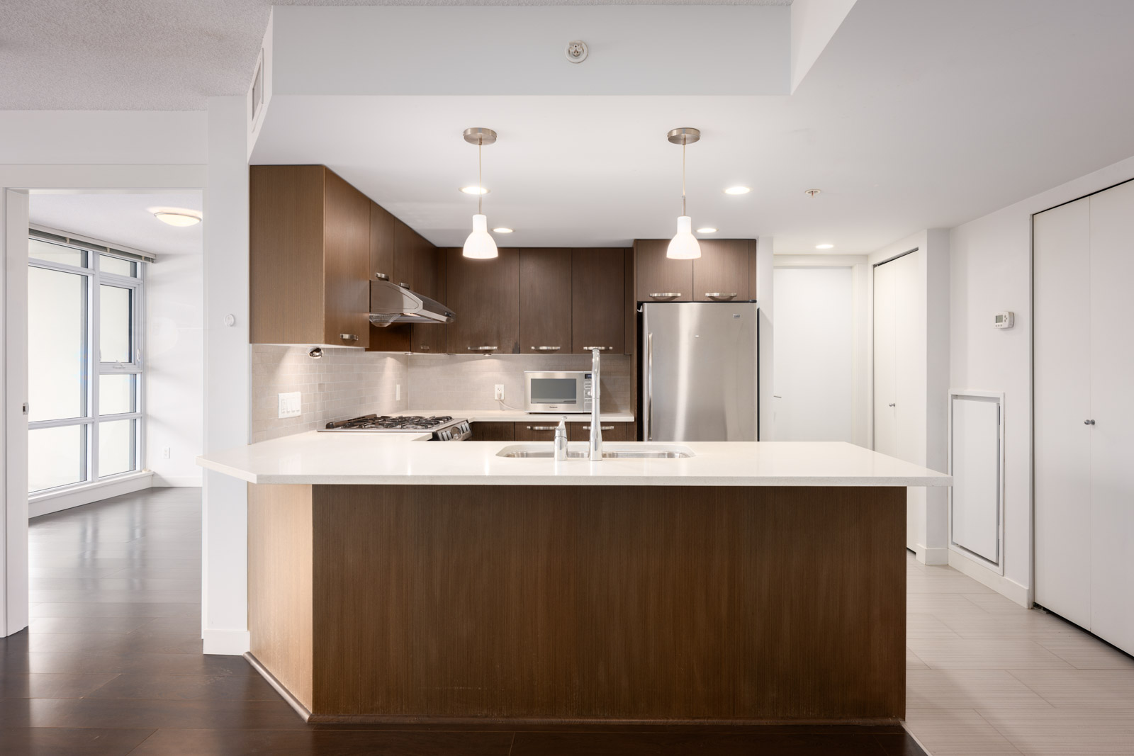 Modern kitchen with wood cabinets, stainless steel appliances, white countertop island, two pendant lights, and an adjacent open living space with large windows.