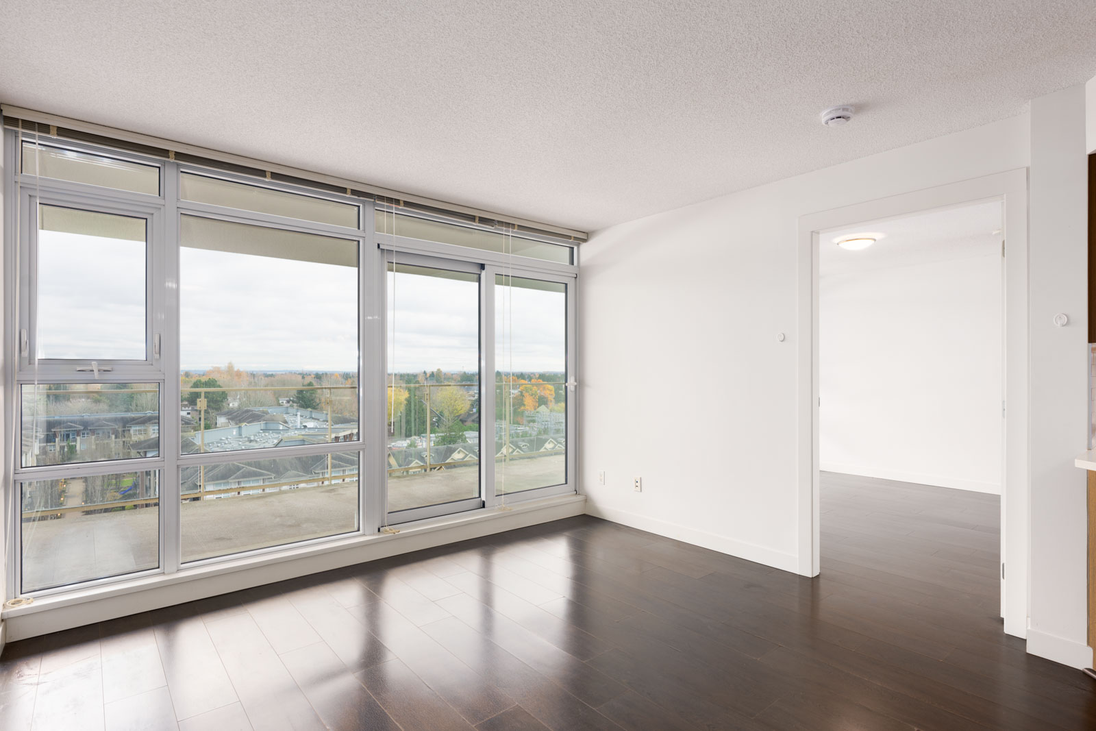 Unfurnished apartment room with large floor-to-ceiling windows, dark wood flooring, and a view of a balcony and cityscape. An open doorway leads to another bright room.