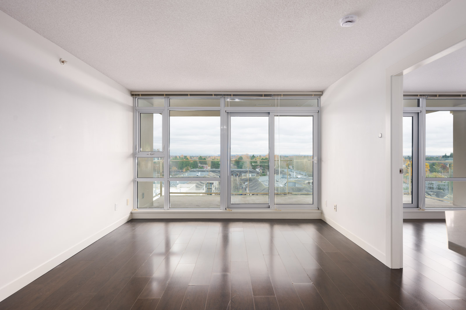 Unfurnished living room with large floor-to-ceiling windows, dark wood flooring, and a view of the cityscape and trees outside.