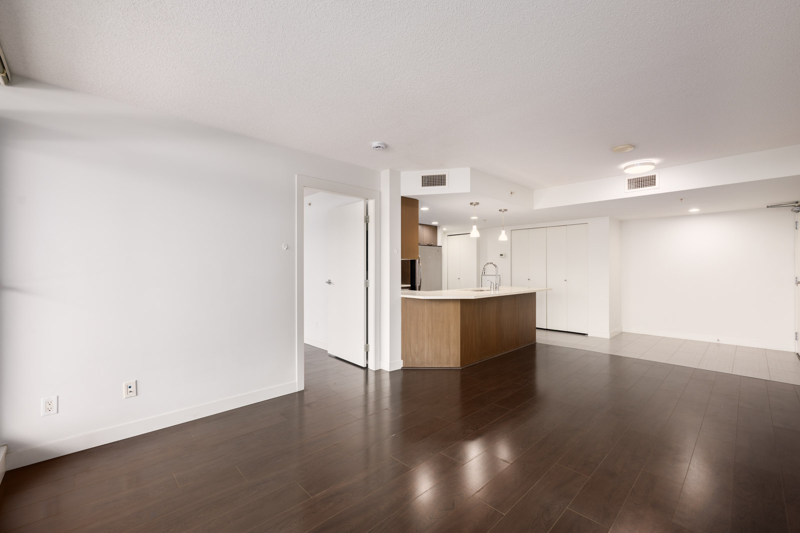 Unfurnished modern apartment interior with dark wood floors, white walls, an open kitchen with an island, and a partially visible door leading to another room.