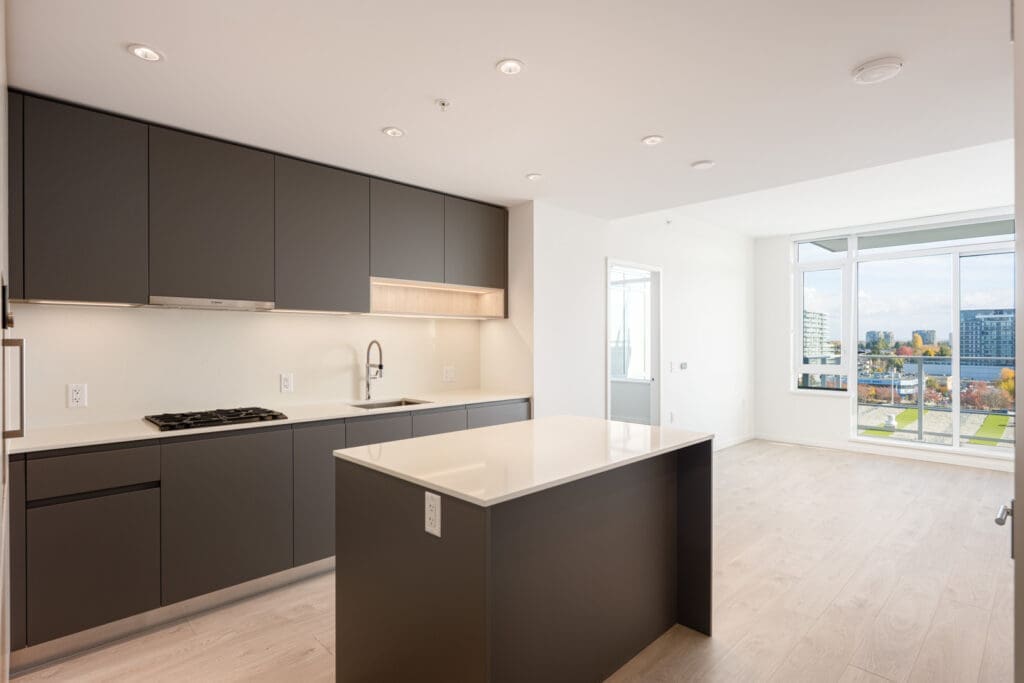 Modern kitchen with matte dark cabinets, gas stove, island, and under-cabinet lighting, adjacent to a bright open living area with large windows and city views.