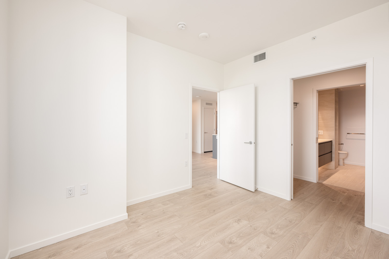 A bright, empty room with light wood flooring, white walls, an open door leading to a hallway, and another door to a bathroom with visible toilet and vanity.