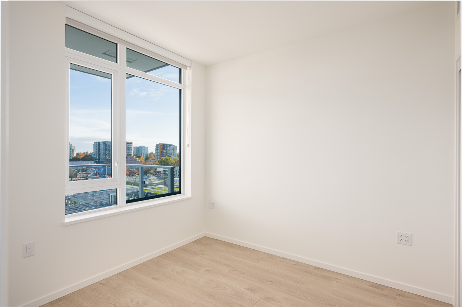 An empty room with light-colored wood flooring, white walls, and a large window showing a cityscape outside.