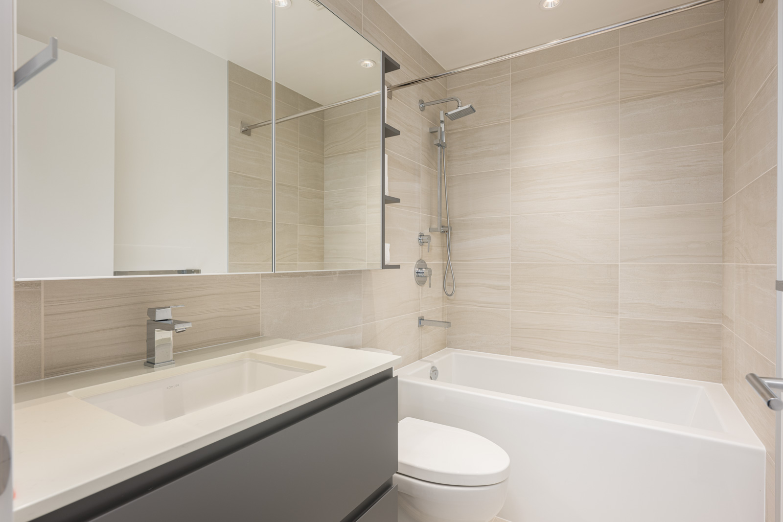 Modern bathroom with beige tiled walls, a white bathtub with a shower, a wall-mounted toilet, a wide vanity with a sink, and a large mirrored cabinet.