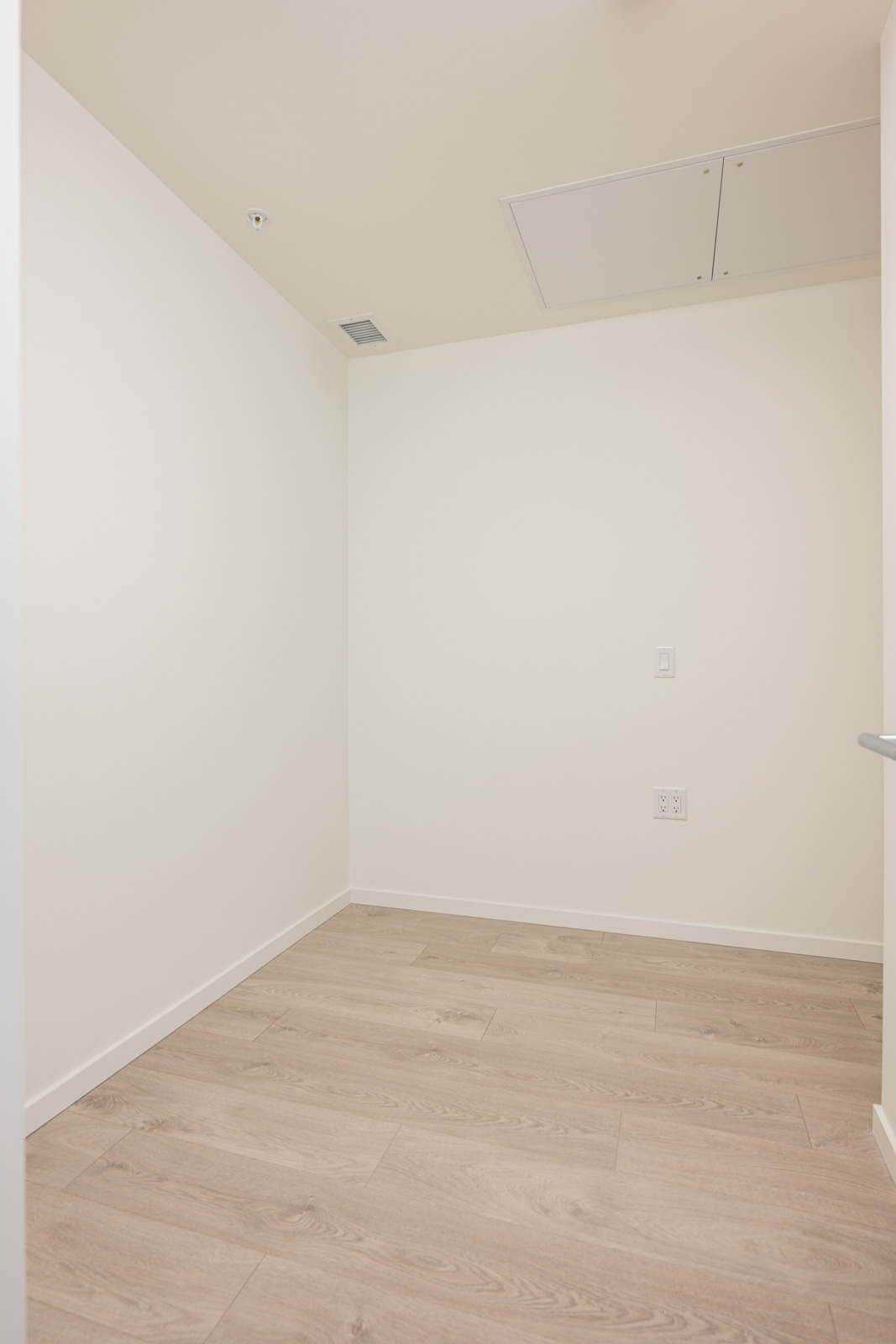 A small empty room with white walls, light wood flooring, a ceiling vent, and visible electrical outlets.