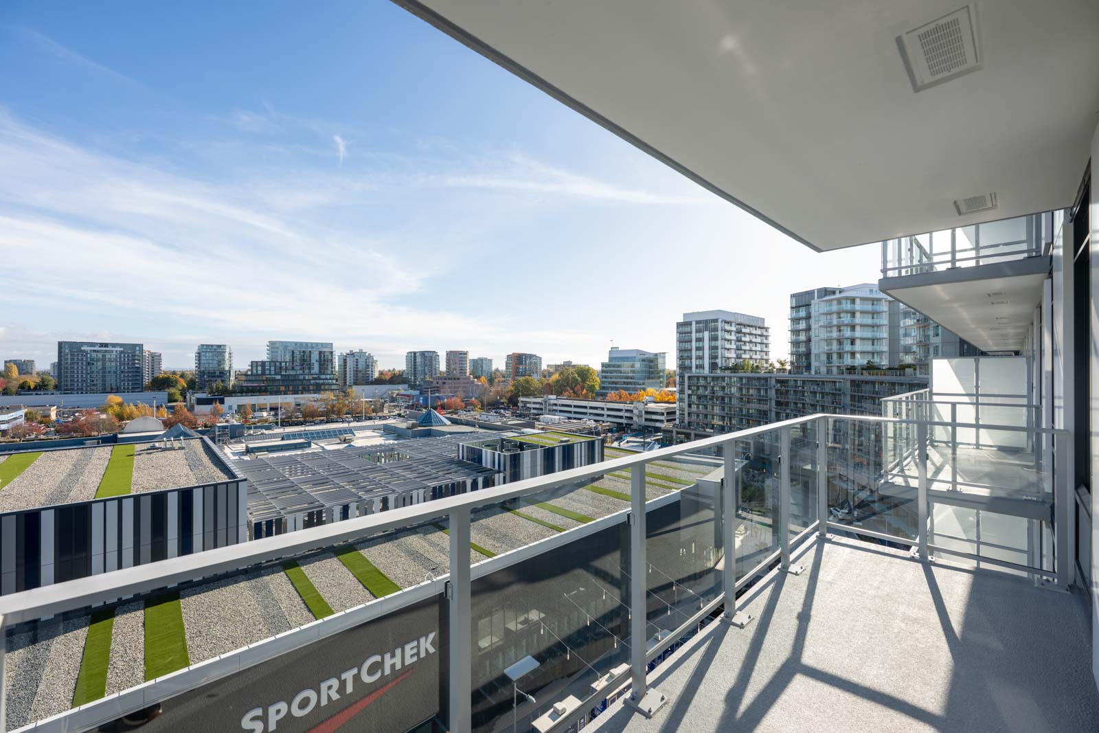 View from a high-rise balcony overlooking a cityscape with modern buildings, parking lots, and a Sport Chek store under a clear blue sky.