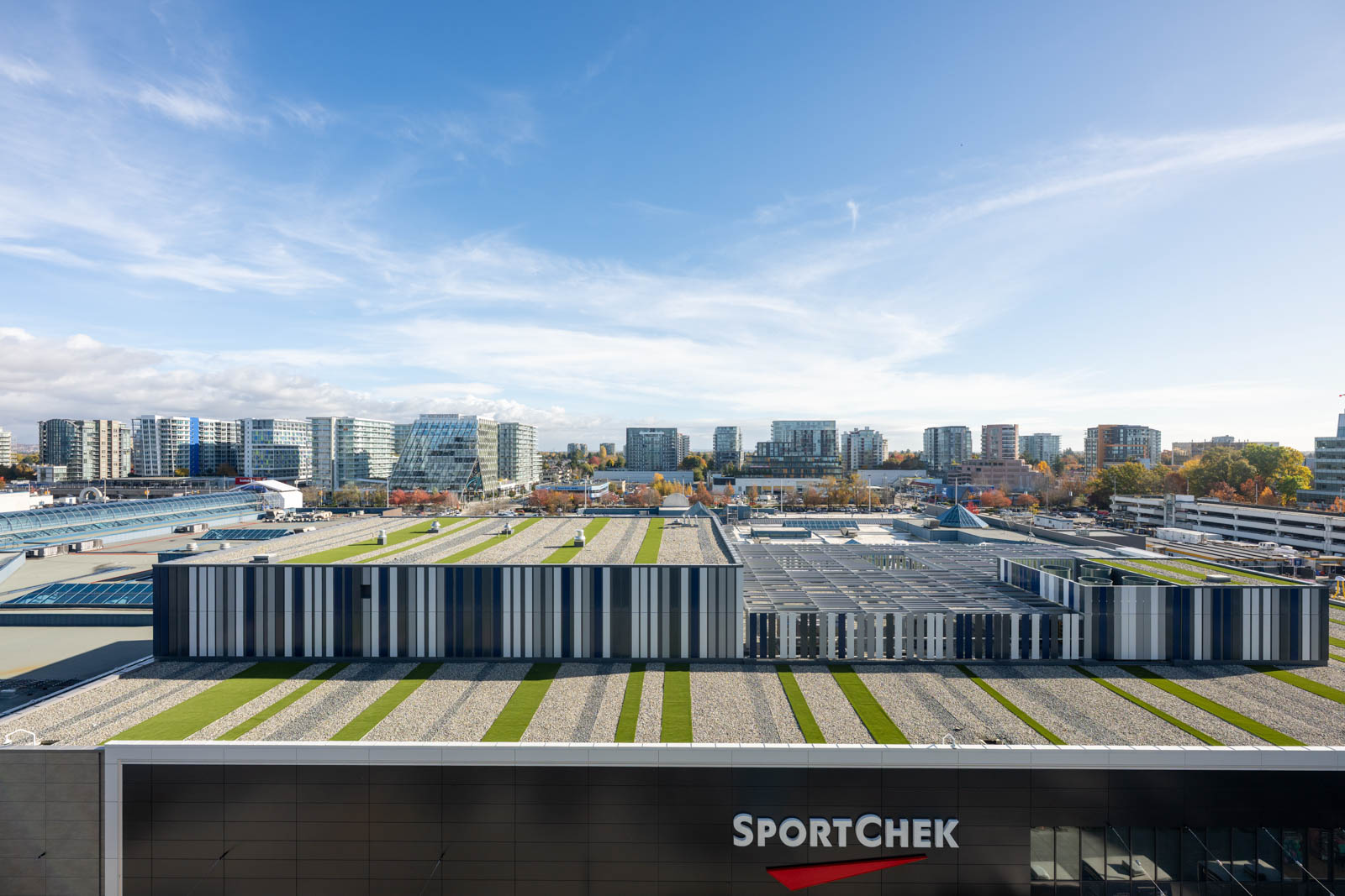 A modern building with a striped facade and green rooftop sections sits in front of a city skyline, with the SportChek logo visible in the foreground.