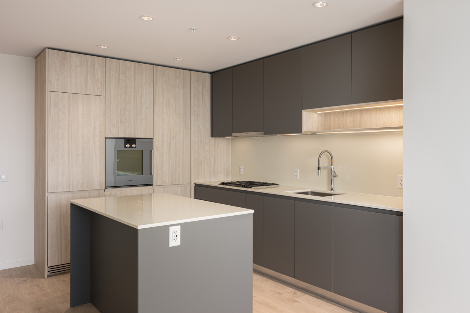 Modern kitchen with matte gray cabinets, built-in oven, gas stove, integrated refrigerator, and a white island countertop under recessed ceiling lights.