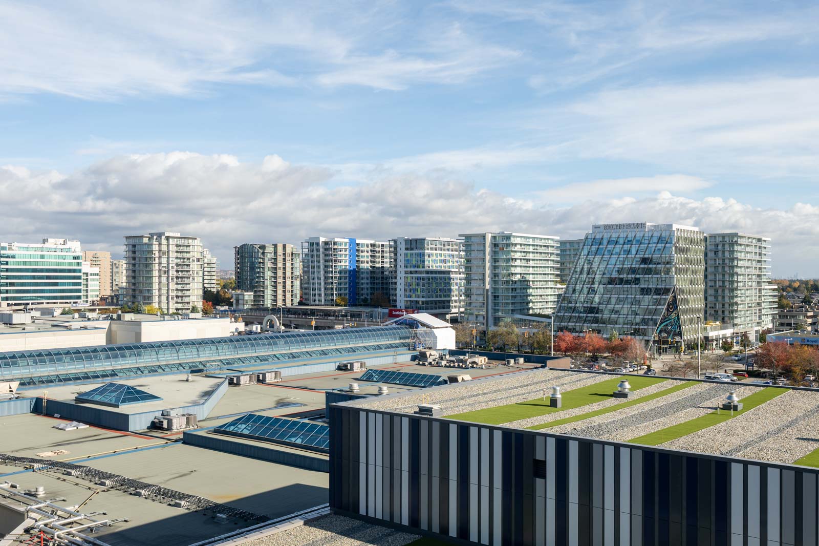 A cityscape featuring modern mid-rise buildings, including a glass-angled office tower, with flat-roofed structures in the foreground under a partly cloudy sky.