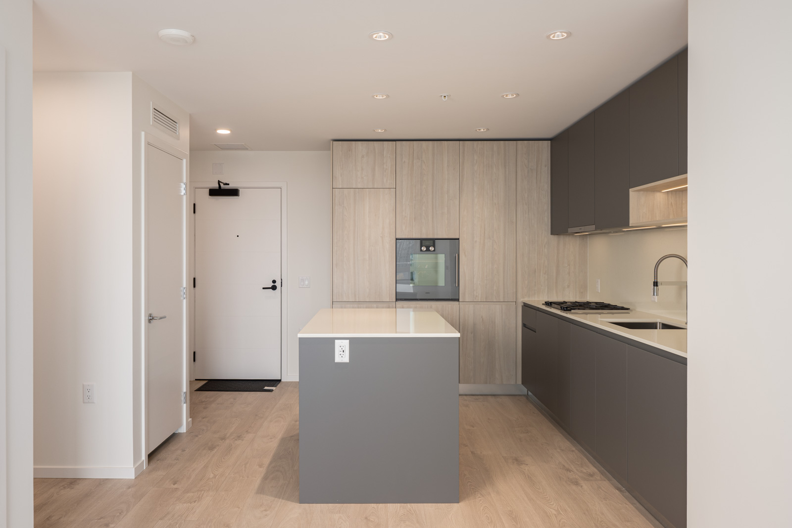Modern kitchen with light wood floors, gray cabinets, a center island, integrated appliances, and a white entrance door in the background.
