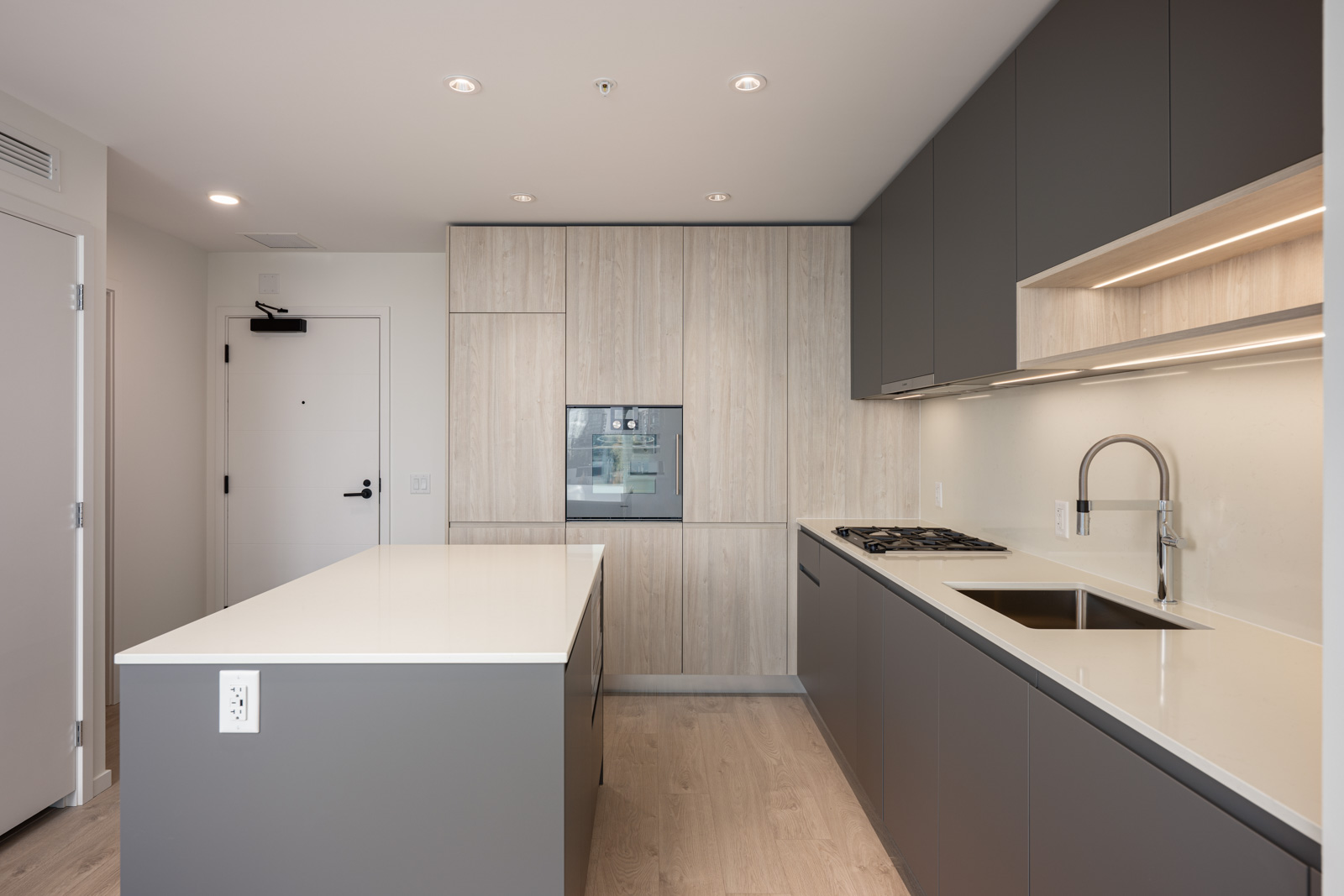 Modern kitchen with gray cabinets, white countertops, built-in oven, gas stove, under-cabinet lighting, and a central island in a bright, minimalist space.