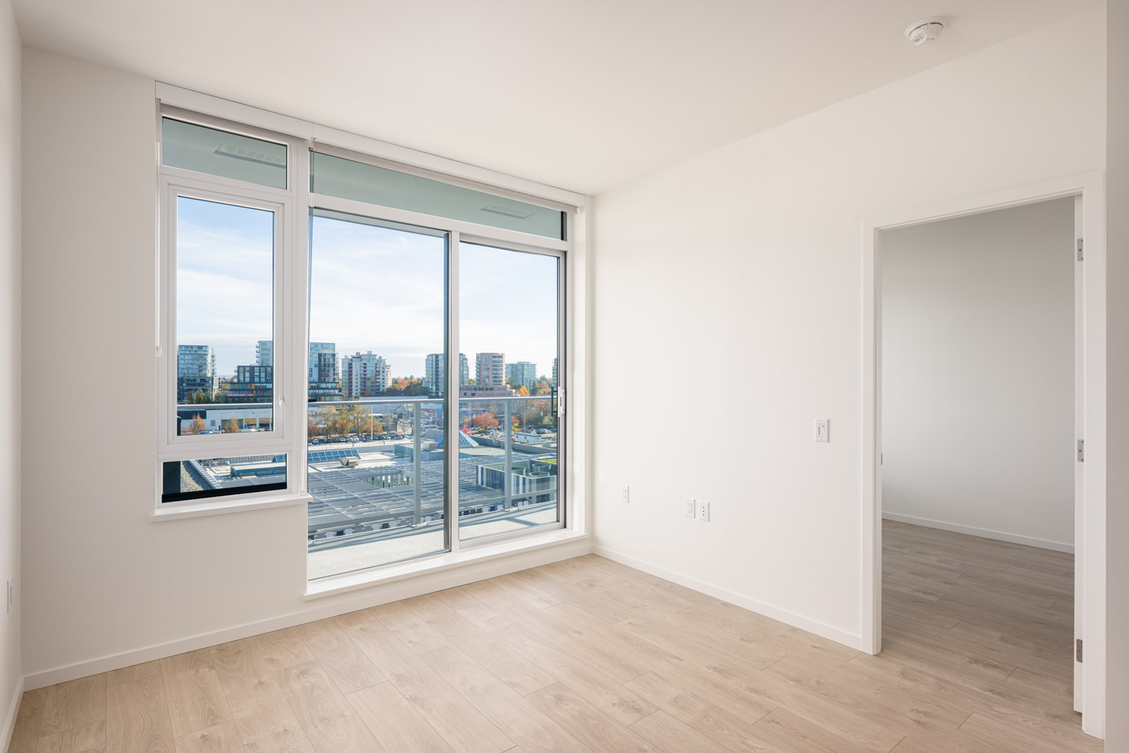 Empty, unfurnished room with light wood flooring, large window and glass door leading to a balcony, and a view of buildings and trees outside. An open doorway leads to another room.