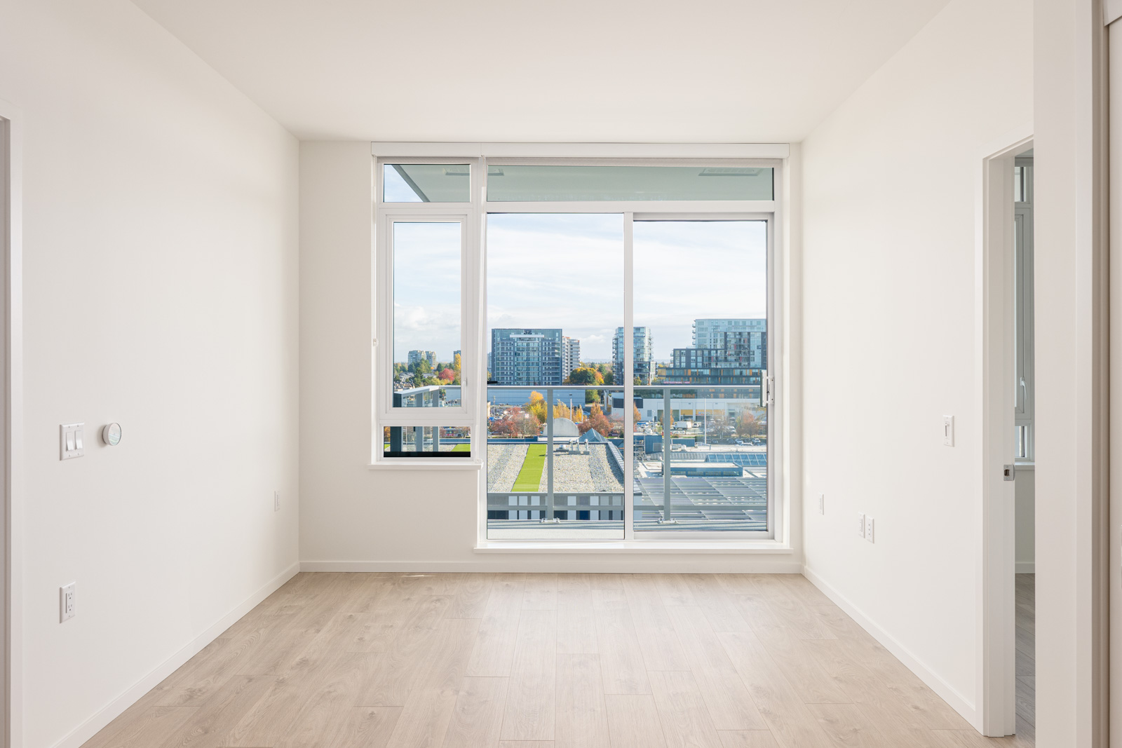 Empty, unfurnished room with light wood flooring, white walls, and large windows overlooking a cityscape with buildings and trees.