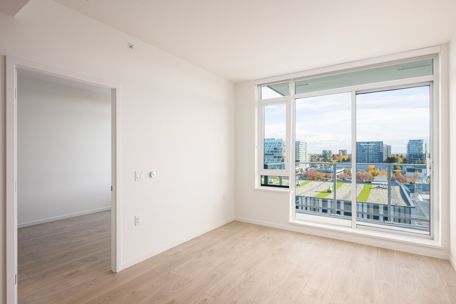 Bright empty room with light wood flooring, large window, glass door to balcony, and a view of modern buildings; adjacent doorway leads to another room.