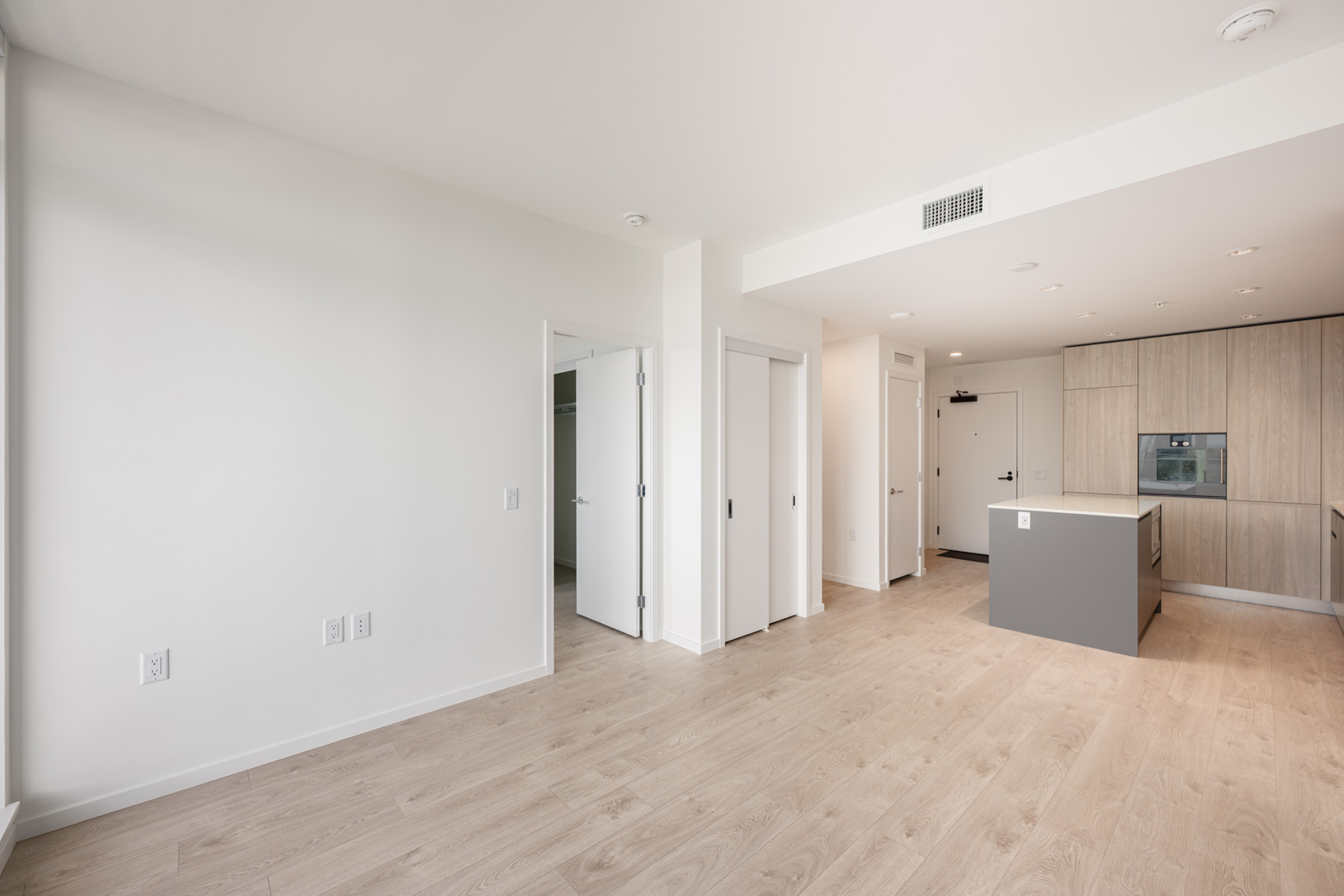 A modern, unfurnished apartment interior with light wood flooring, white walls, a kitchen area with wood cabinets, and multiple white doors.