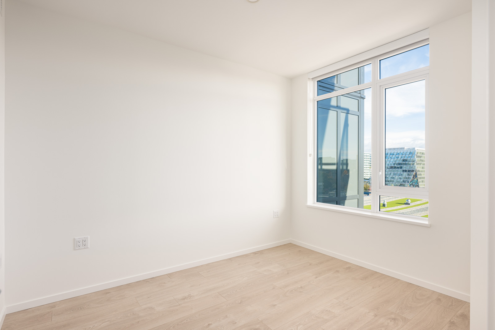 Empty, unfurnished room with white walls, light wood flooring, and a large window showing modern buildings and a clear sky outside.