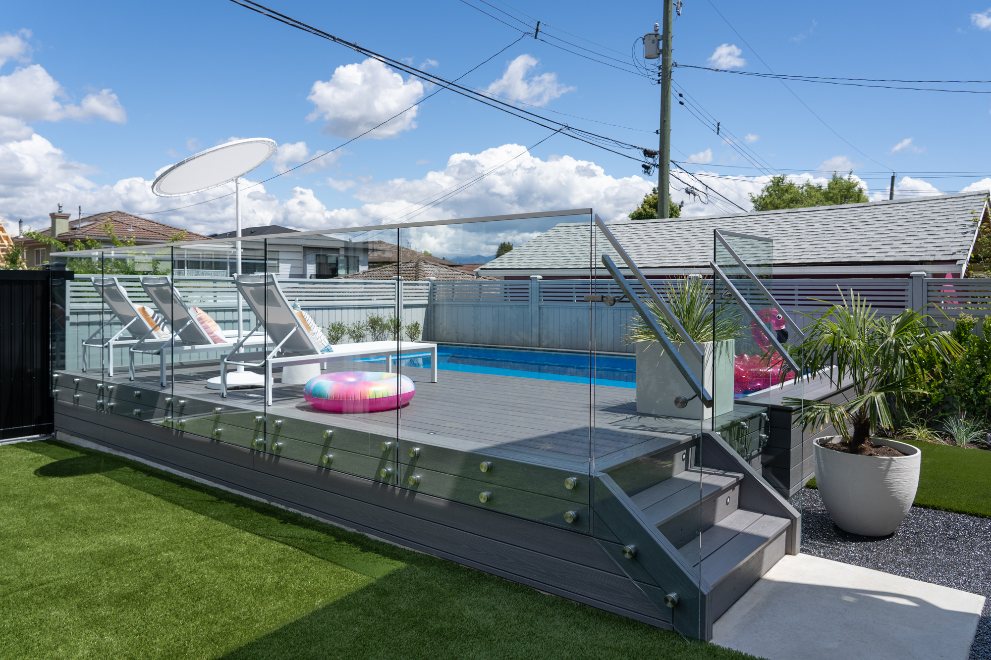 A modern backyard pool area with glass fencing, two lounge chairs, a white umbrella, and an inflatable float, set on a raised deck beside artificial grass.