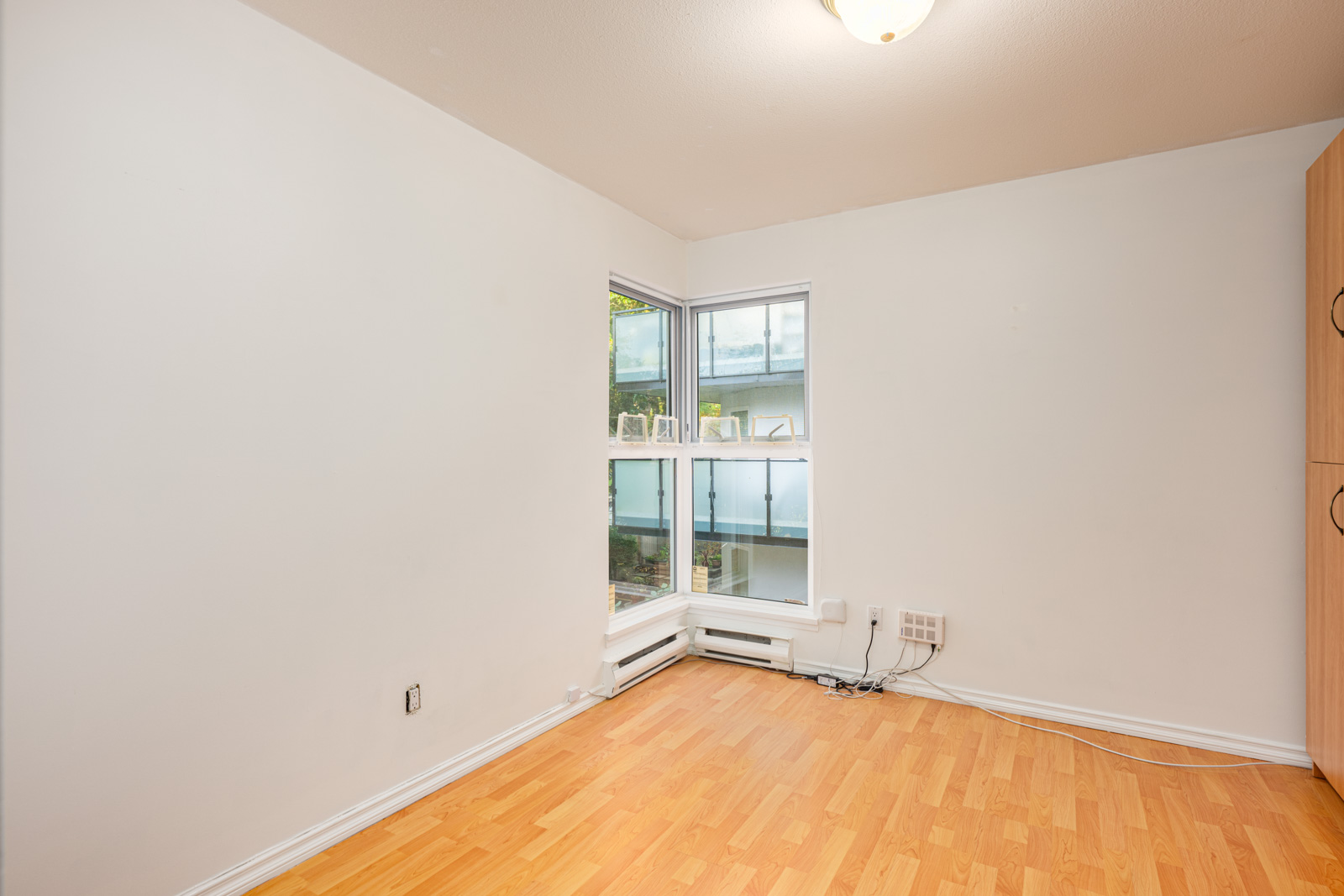 Unfurnished room with light wood flooring, white walls, a corner window with small plants on the sill, and visible electrical outlets and cables along the floor.