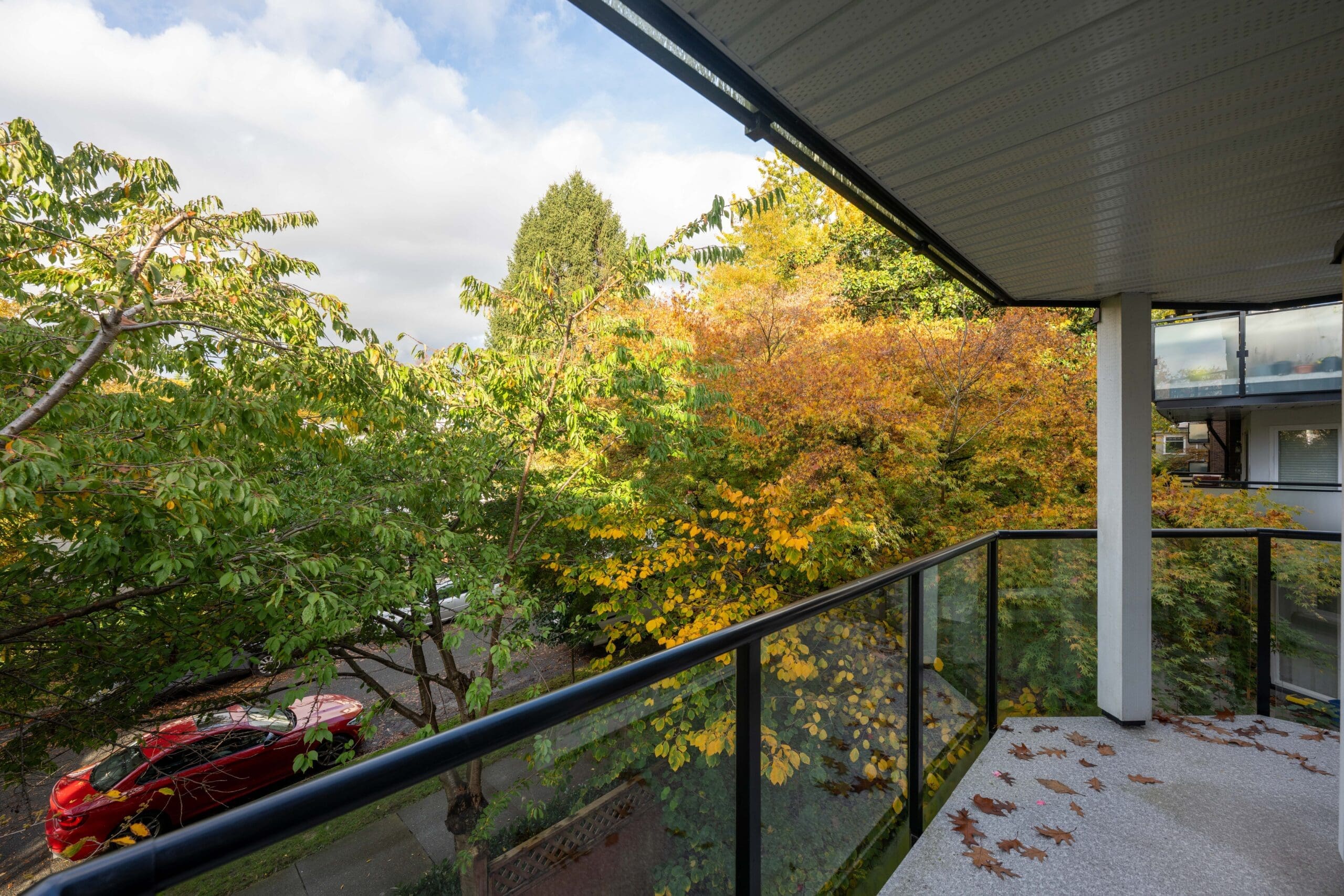 A balcony with glass railing overlooks trees with green and yellow leaves. A red car is parked on the street below. Some leaves are scattered on the balcony floor.