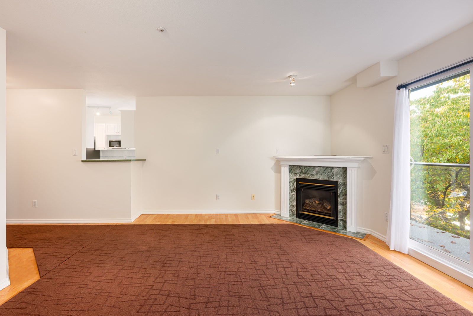 Unfurnished living room with a brown carpet, fireplace, sliding glass door to a balcony, and view into a white kitchen.