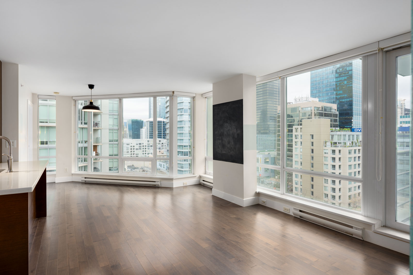 Modern empty apartment with large floor-to-ceiling windows, hardwood floors, and cityscape views; kitchen area visible on the left.