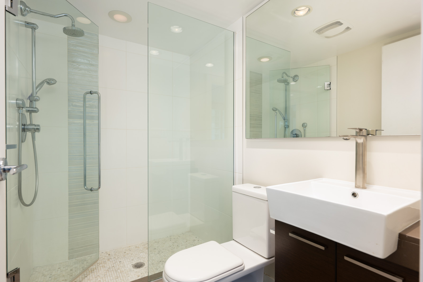Modern bathroom with glass-enclosed shower, white toilet, square sink, brown vanity, and wall mirror under recessed lighting.