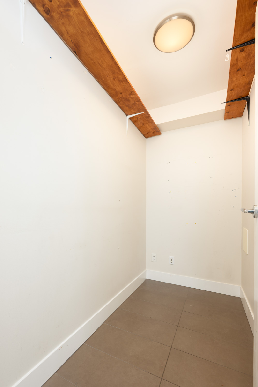 Empty, narrow room with white walls, brown ceiling beams, tiled floor, and a round ceiling light. A few wall-mounted brackets and hooks are visible.