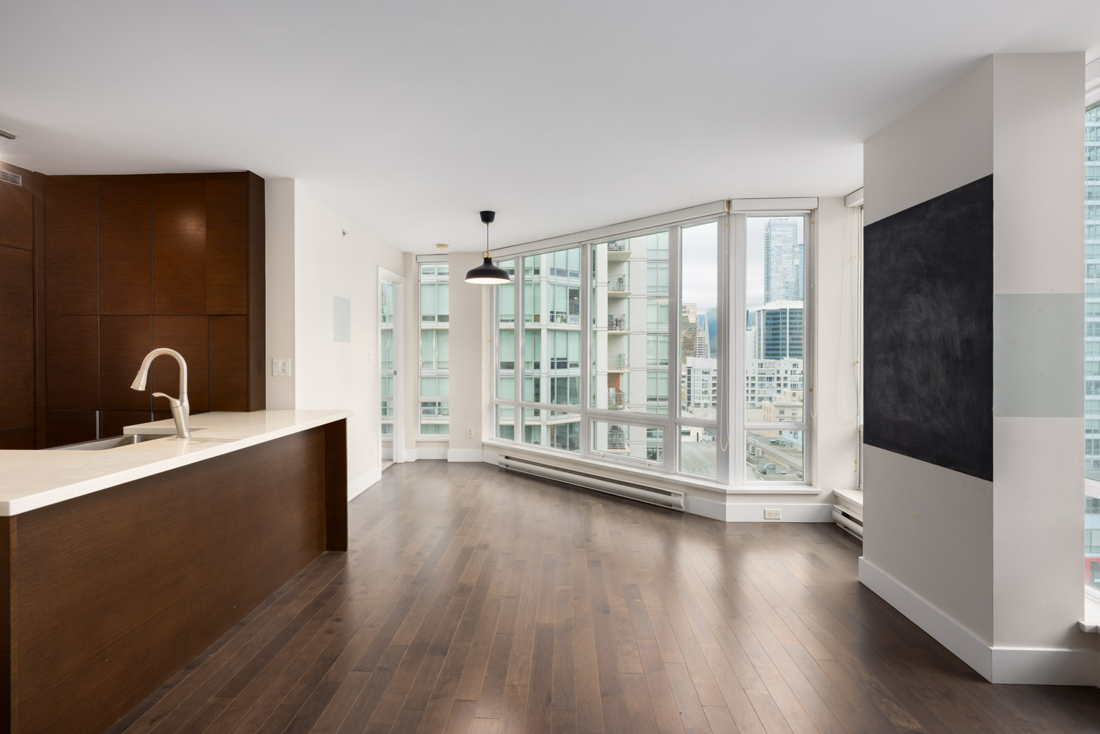 Modern, unfurnished apartment with dark wood floors, large windows offering city views, a kitchen island with sink, and a blank wall chalkboard.