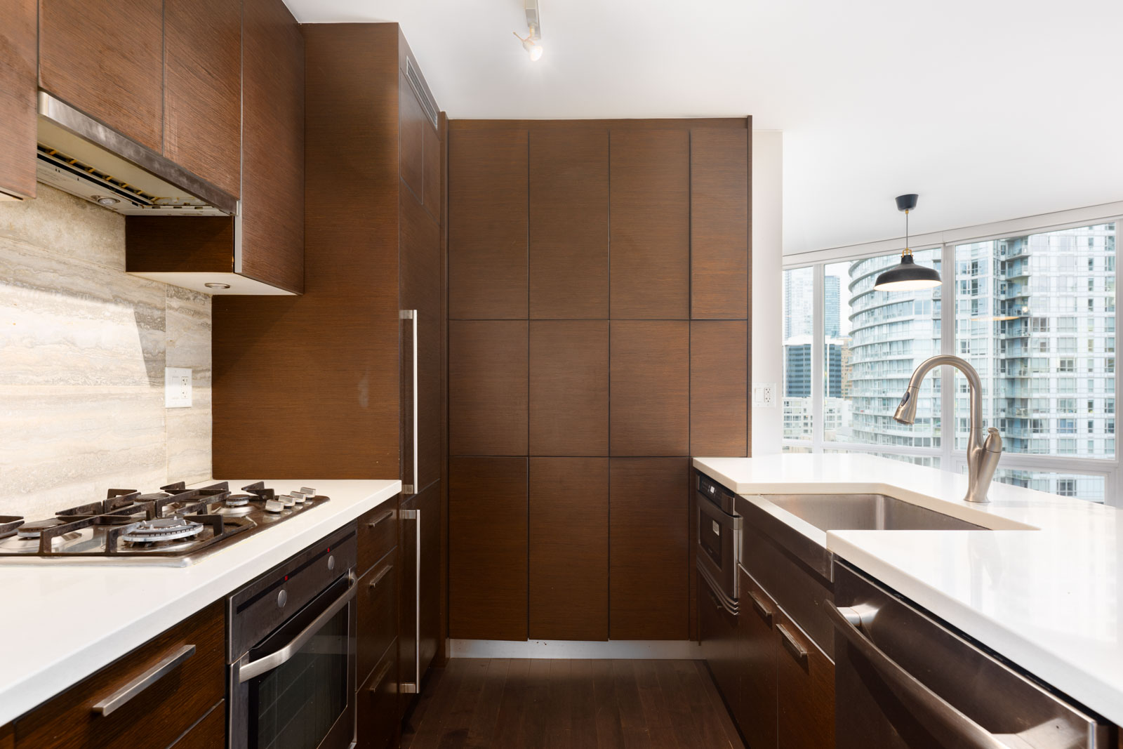 Modern kitchen with dark wood cabinets, white countertops, built-in appliances, gas stove, and large windows overlooking city buildings.