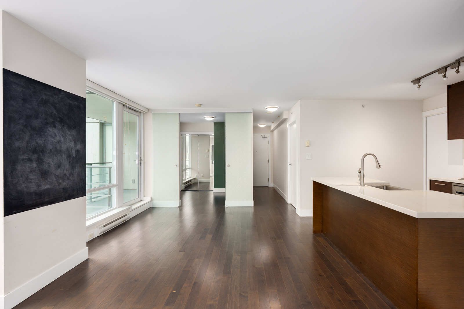 Modern empty apartment with dark wood floors, an open kitchen with an island, large windows, and a balcony door. White walls and ceiling lights provide a bright, clean atmosphere.