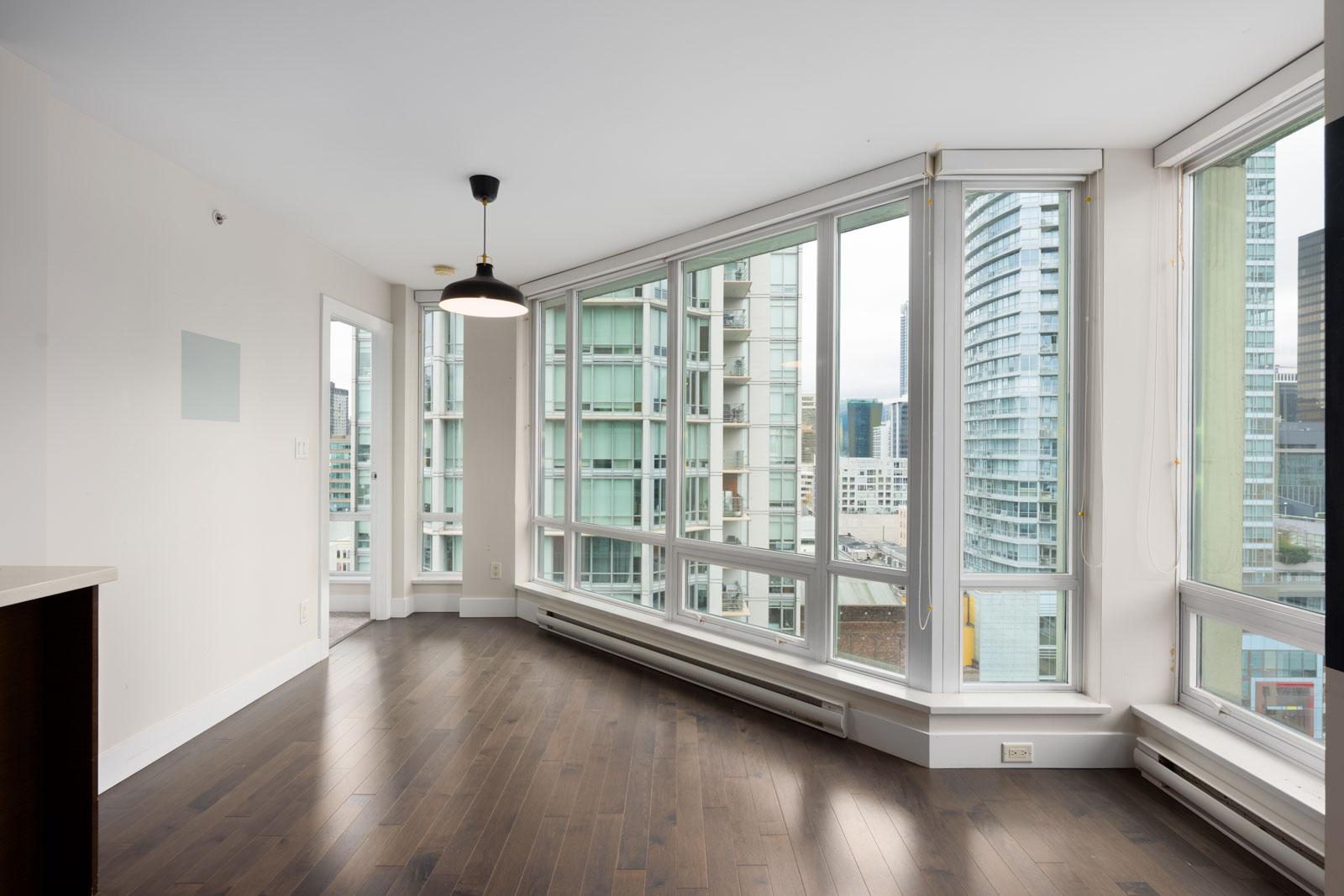 Unfurnished apartment room with large floor-to-ceiling windows, city views, dark wood flooring, white walls, and a ceiling light fixture.