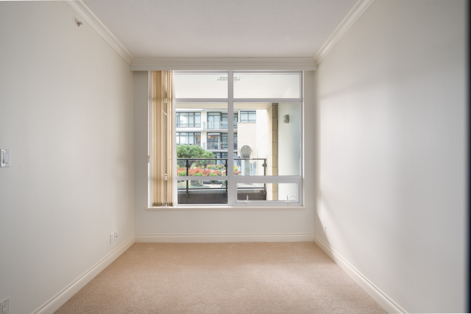 Empty room with beige carpet and off-white walls, featuring a large window overlooking a balcony and a modern apartment building in the background.