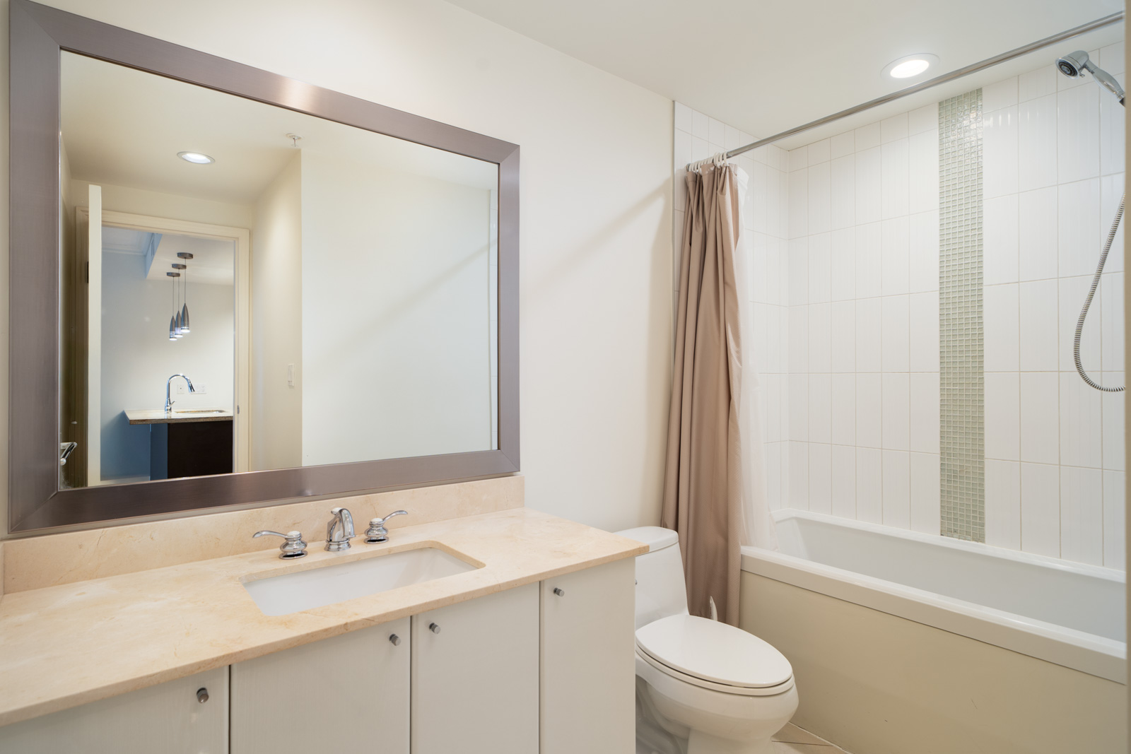 Modern bathroom with a large mirror, beige countertop, white sink, toilet, and a bathtub with a shower curtain and tiled wall. Recessed lighting brightens the space.