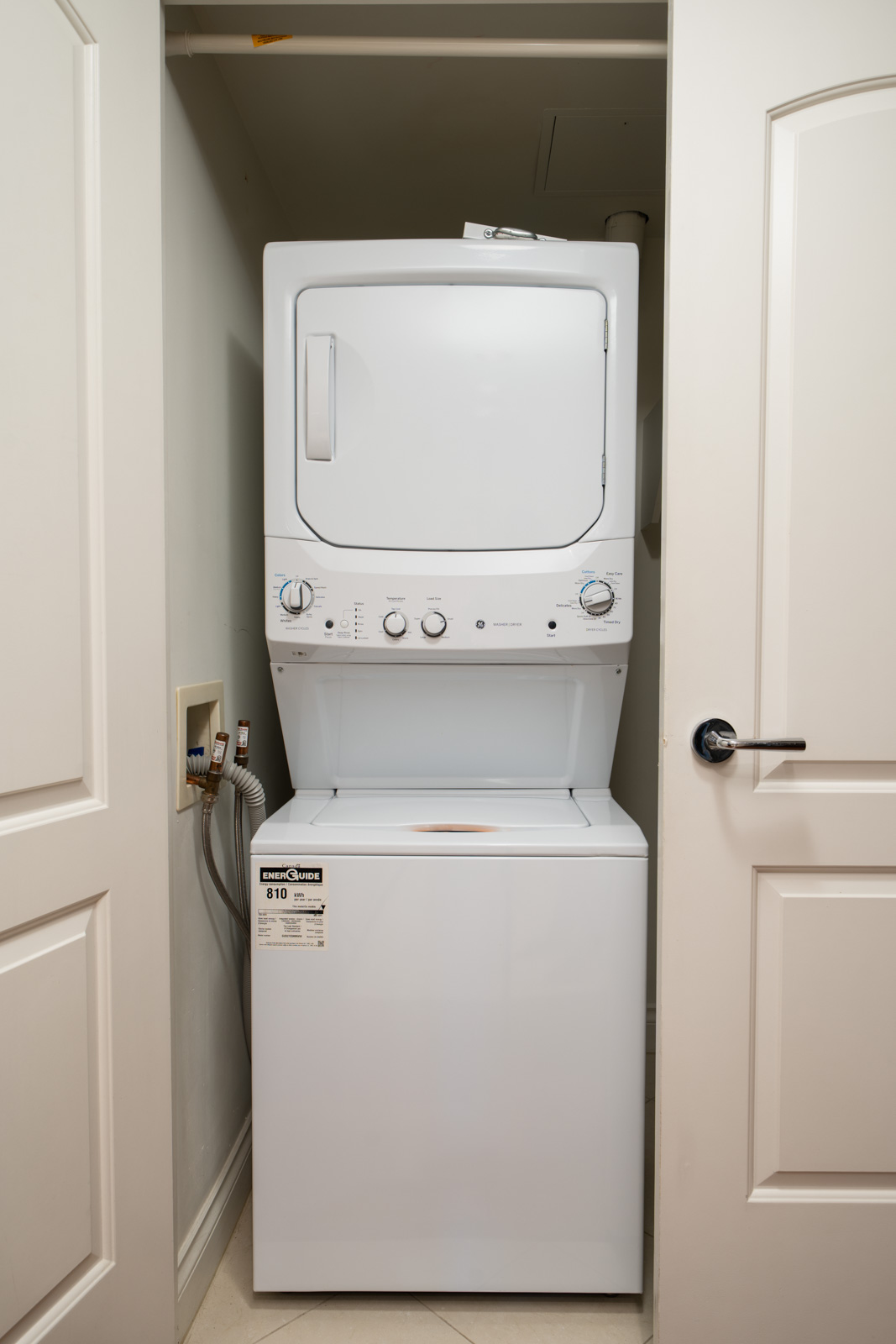 A white stacked washer and dryer unit is installed in a small laundry closet with white doors partially open.