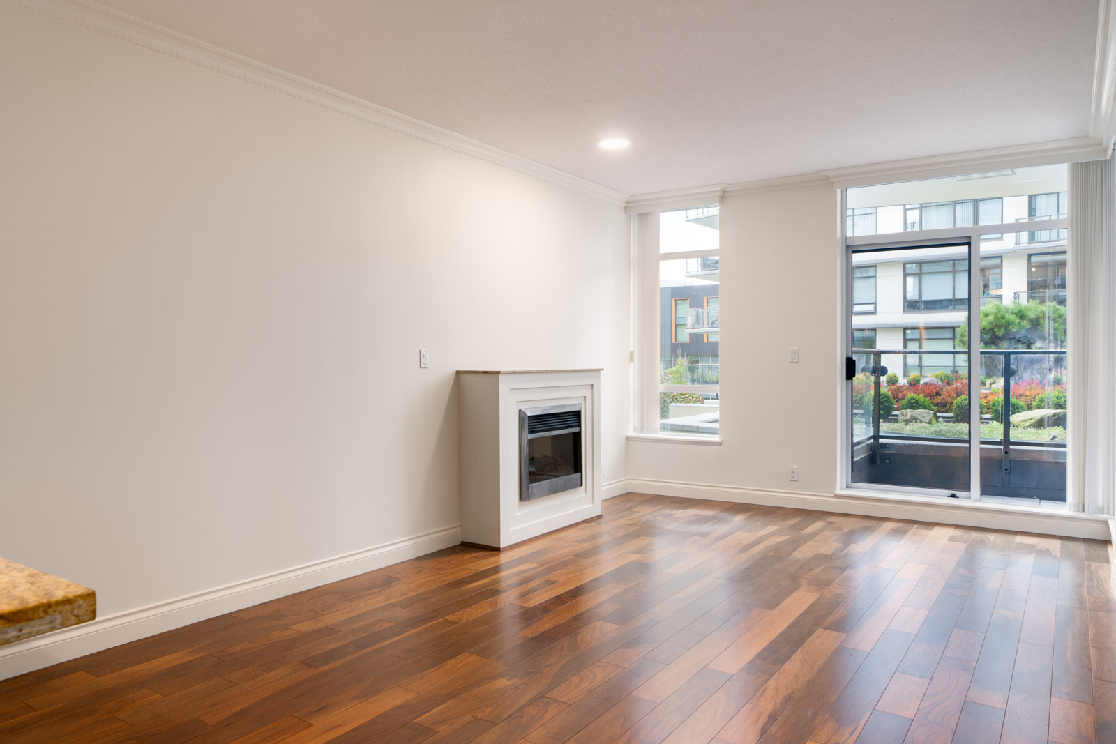 Unfurnished living room with hardwood floors, a white fireplace, large windows, and a glass door leading to a balcony with a view of an apartment complex and landscaped area.