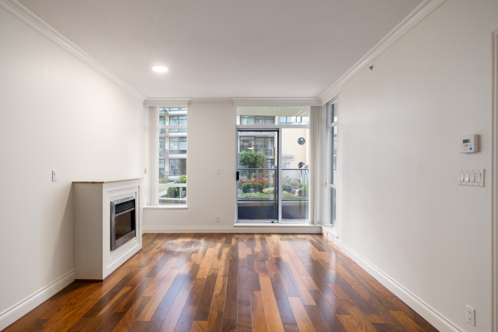 Empty living room with hardwood floors, white walls, a fireplace on the left, large windows, and a glass door leading to a balcony with a garden view.
