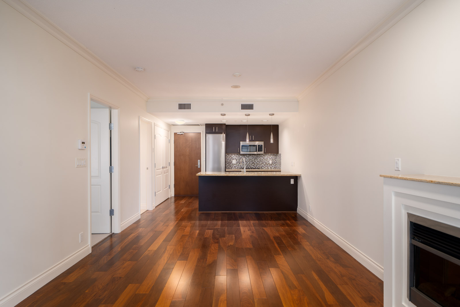 Unfurnished apartment interior with hardwood floors, white walls, a kitchen with dark cabinets and stainless steel appliances, and a fireplace on the right.