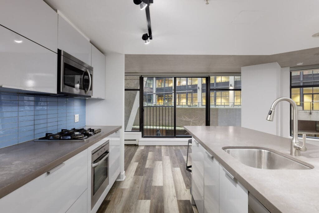 Modern kitchen with white cabinets, gray countertops, gas stove, built-in microwave, blue tile backsplash, island sink, and large windows overlooking a city building.