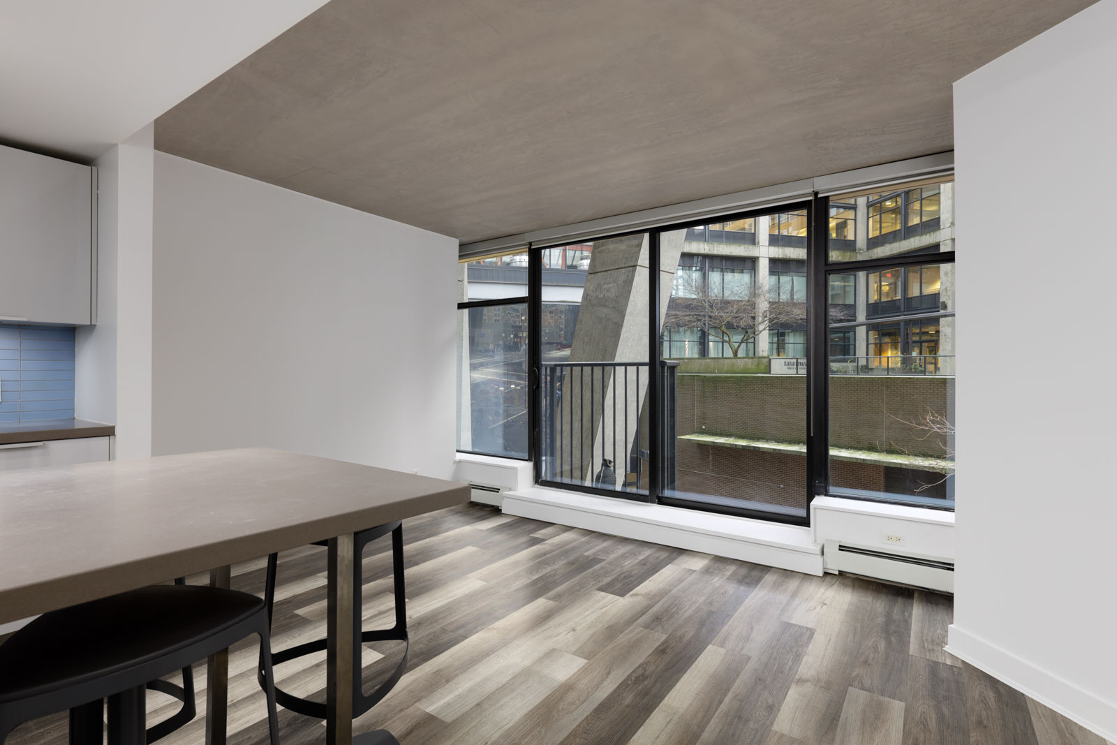 Modern apartment interior with large floor-to-ceiling windows, light wood flooring, white walls, and a view of an urban building exterior.