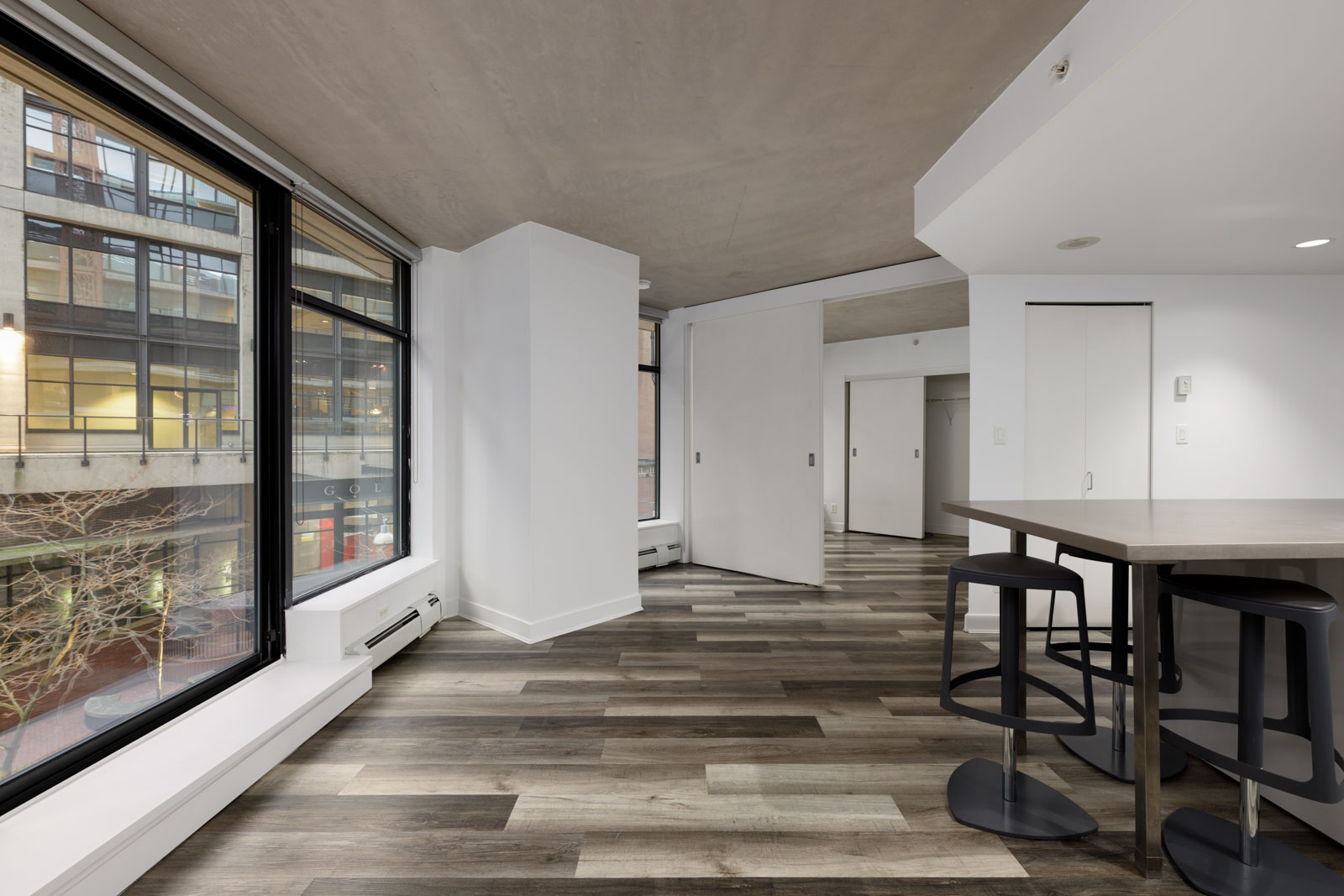 Modern apartment interior with large windows, wood-patterned flooring, white walls, a kitchen island, and two black bar stools; exterior building visible through windows.