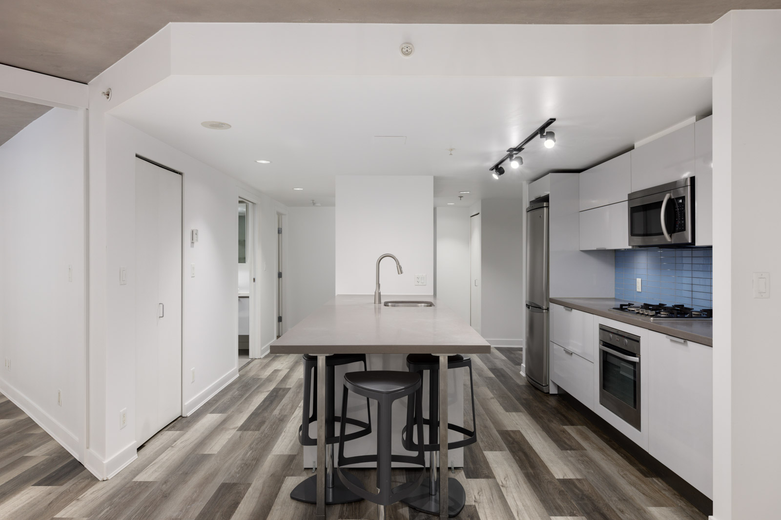 Modern kitchen with an island and bar stools, stainless steel appliances, white cabinets, blue tile backsplash, and wood-look flooring.
