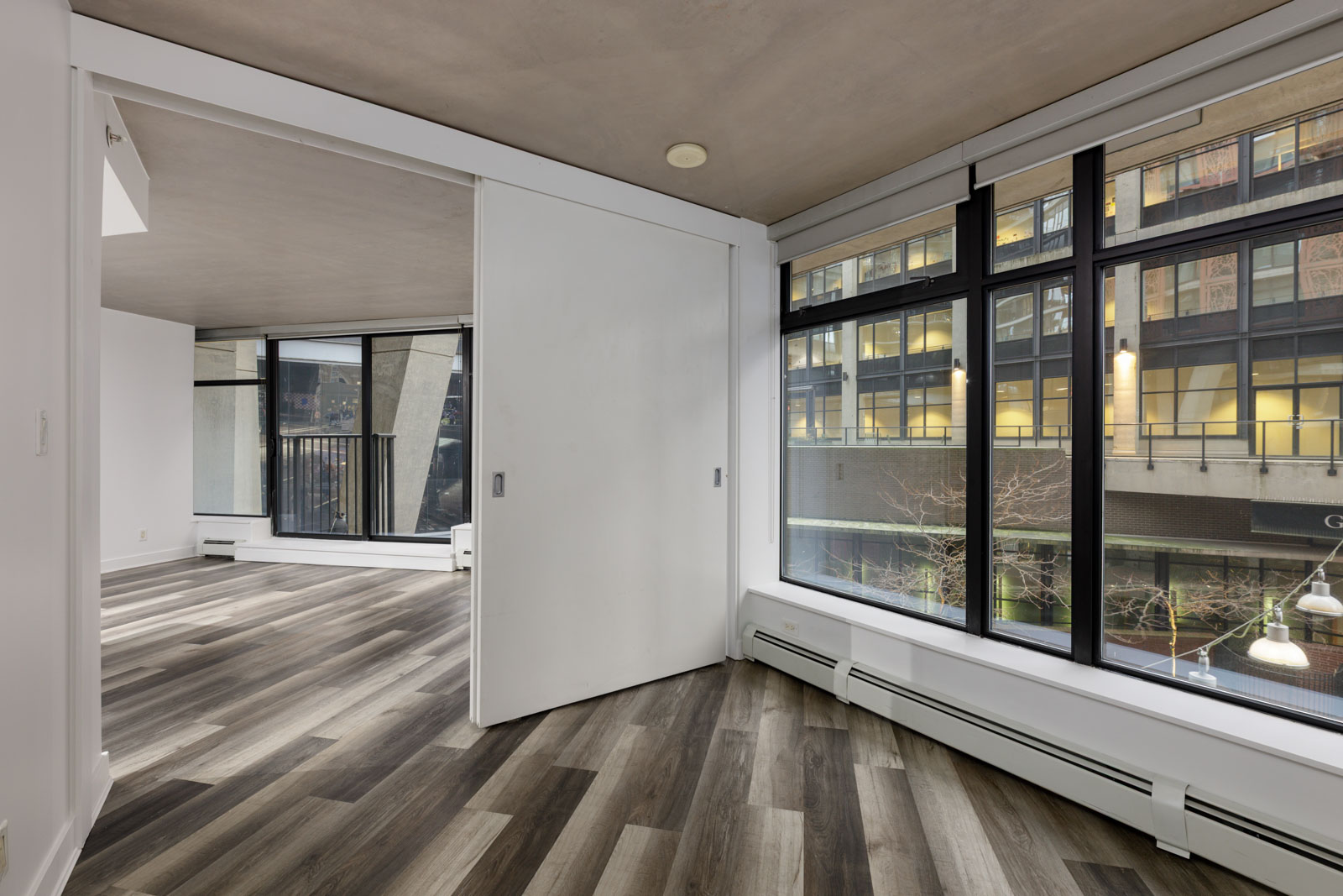 Modern apartment with large windows, gray wood flooring, and sliding doors; view of city building and streetlights outside.
