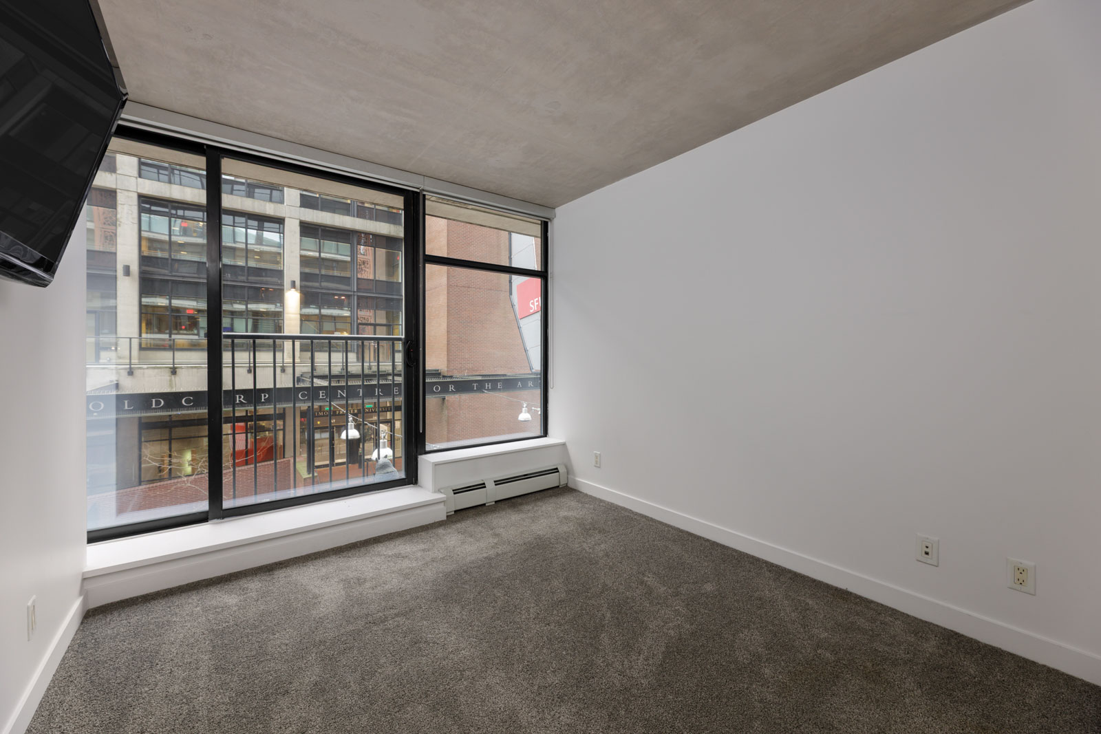 Unfurnished room with gray carpet, white walls, large floor-to-ceiling window, and a view of a building across the street. Wall-mounted TV and baseboard heater visible.