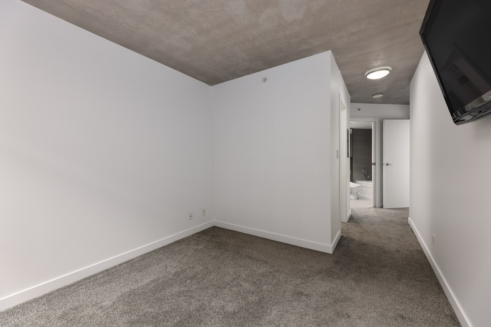 Empty room with white walls, gray carpet, and concrete ceiling; a wall-mounted TV is on the right, and an open door leads to a bathroom in the background.