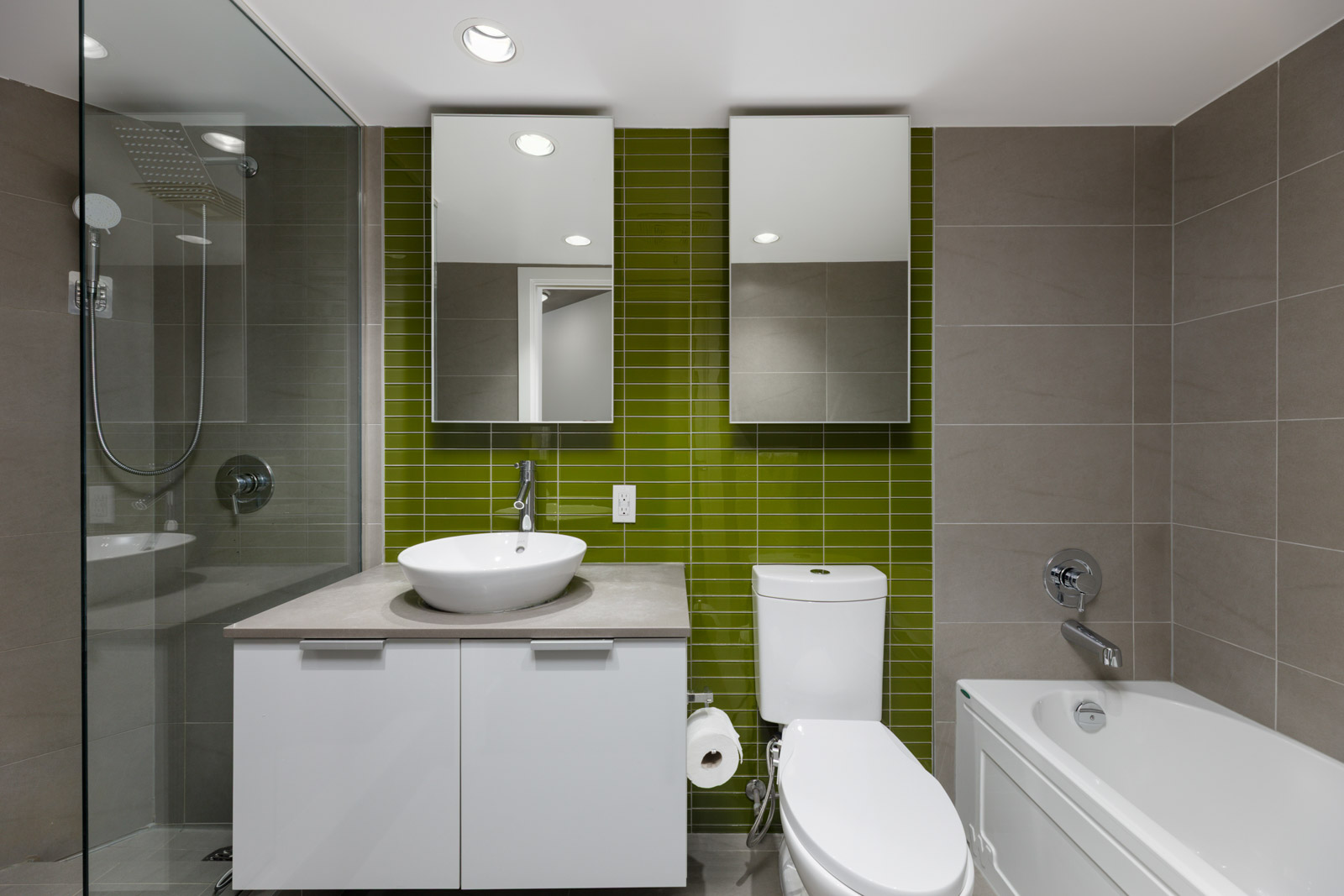 Modern bathroom with a green tile accent wall, white sink and vanity, wall mirrors, toilet, bathtub, and a glass-enclosed shower.