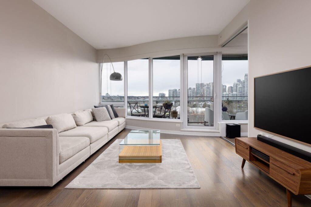 Modern living room with a beige sofa, glass coffee table, TV on a wooden stand, and large windows showing a city skyline and water view.