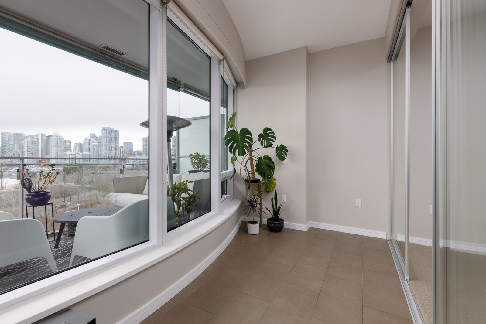 A bright indoor space with large windows, beige walls, potted plants, and a view of a city skyline and patio with white chairs.