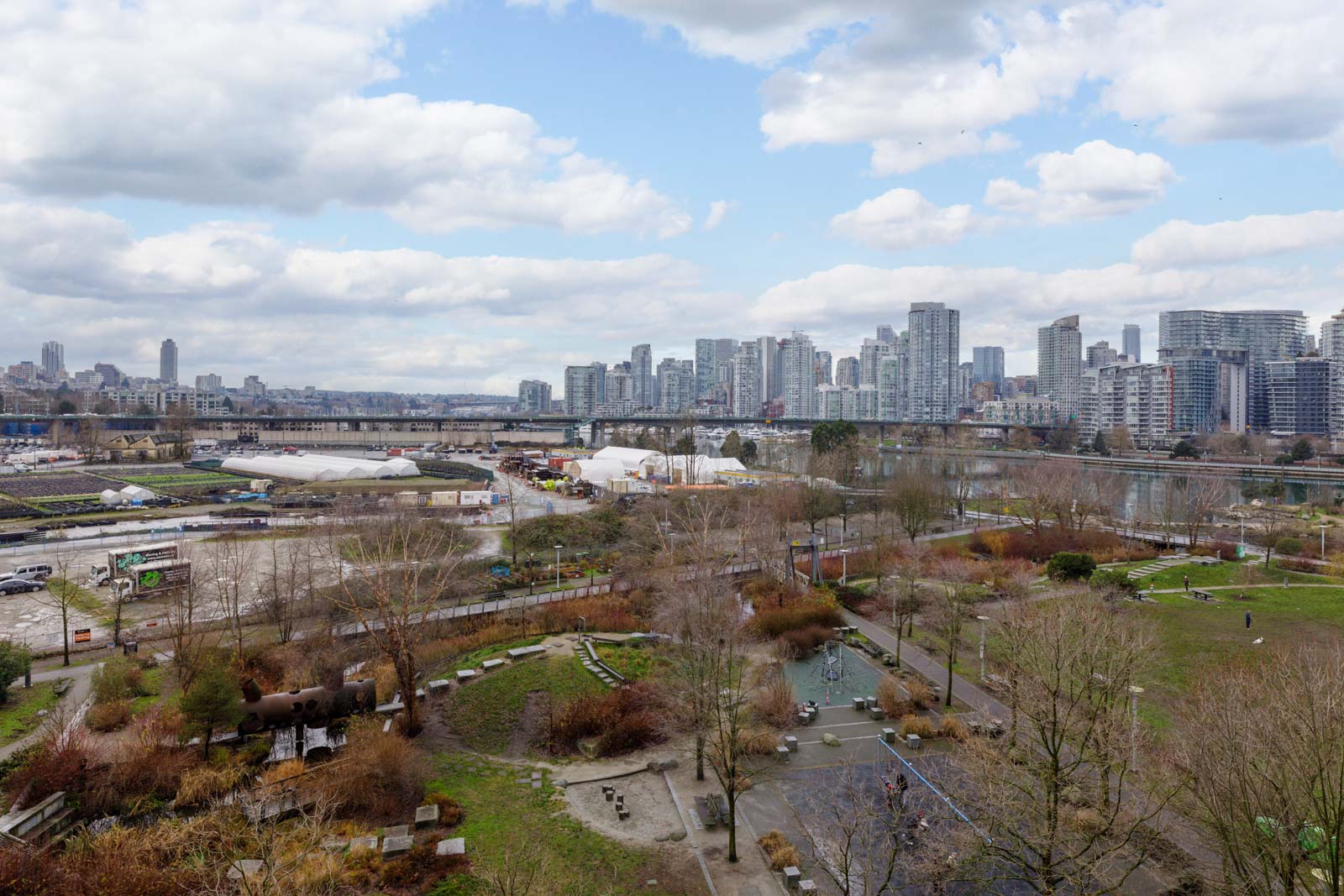 A cityscape view featuring high-rise buildings, a railway bridge, tents, and a park with trees and walking paths under a partly cloudy sky.